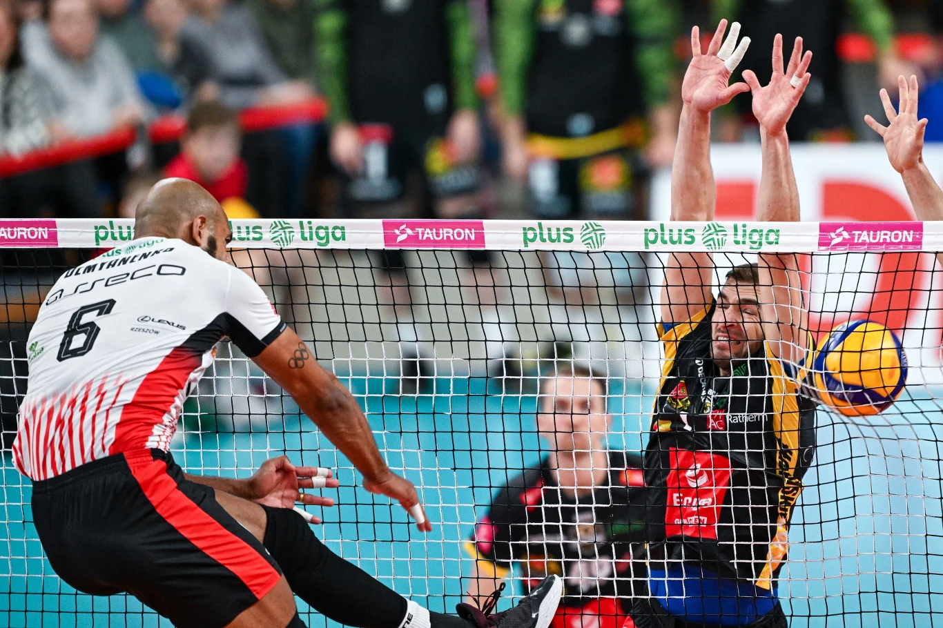 Dynamiczna akcja podczas meczu siatkarskiego, zawodnik w białym stroju atakuje piłkę przy siatce, podczas gdy rywal z drużyny przeciwnej próbuje go zablokować z wyskoku.