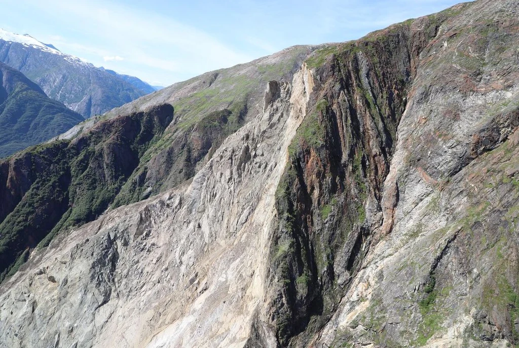 Strome zbocza górskie z widocznymi przepaściami, kontrastujące zielone fragmenty roślinności z surową, skalistą powierzchnią i jasnym niebem w tle
