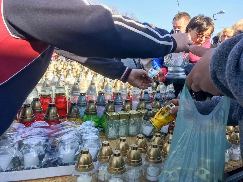 Ludzie wybierają znicze na stoisku z różnokolorowymi lampionami nagrobnymi ustawionymi w rzędach, ręce klientów sięgają po wybrane znicze, a niektóre osoby wkładają je do plastikowych toreb.