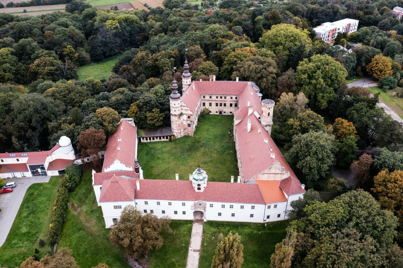 Zamek z czerwonym dachem otoczony gęstym lasem, z centralnym dziedzińcem i charakterystycznymi basztami, widoczny z lotu ptaka w otoczeniu zielonych drzew oraz budynków gospodarczych.