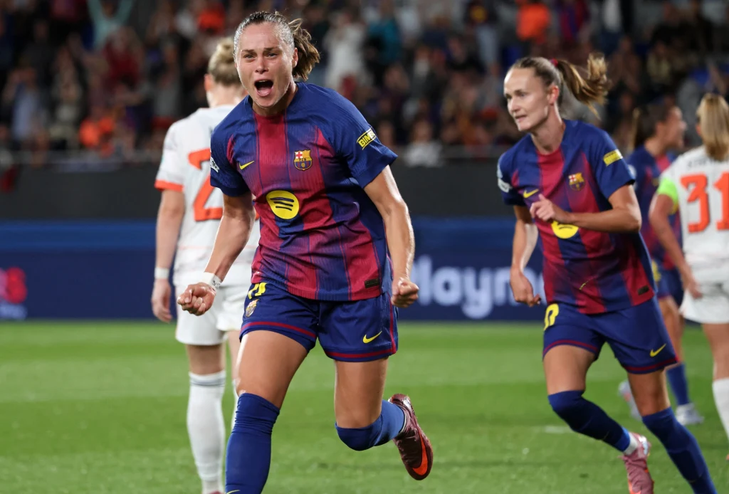 Zawodniczka drużyny FC Barcelona świętuje zdobycie gola na boisku piłkarskim, a w tle widać jej drużynową koleżankę i przeciwniczki w pomarańczowych strojach, trybuny pełne kibiców.