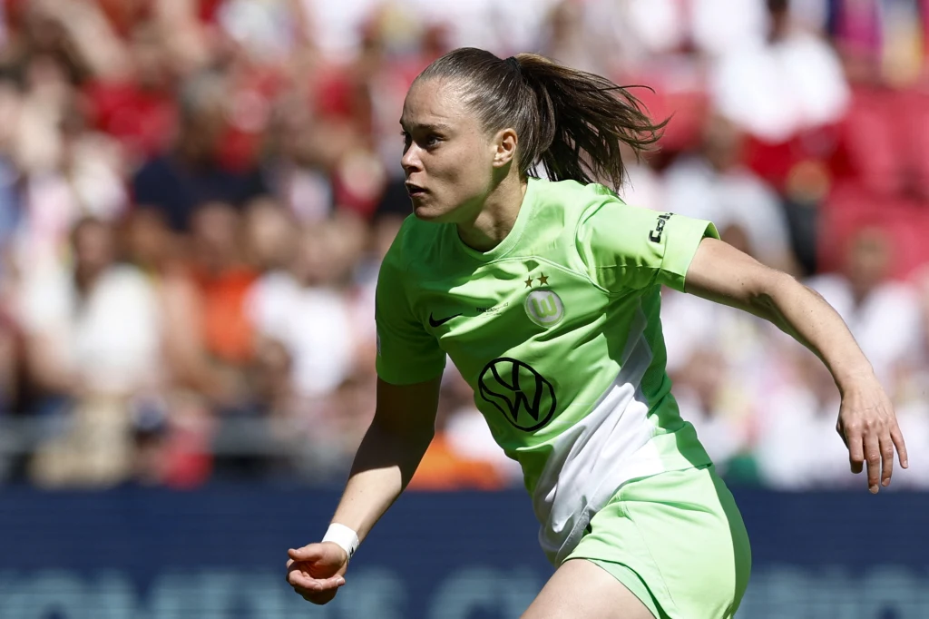 Ewa Pajor w barwach Wolfsburga Kobieta ubrana w zielony strój piłkarski biegnie po murawie na stadionie podczas meczu piłki nożnej, w tle widoczne rozmyte sylwetki kibiców.