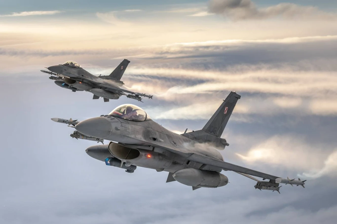 Dwa polskie samoloty myśliwskie F-16 lecące w szyku nad chmurami na wysokości, przy zachmurzonym niebie. Maszyny wojskowe wyraźnie oznaczone narodowymi znakami.