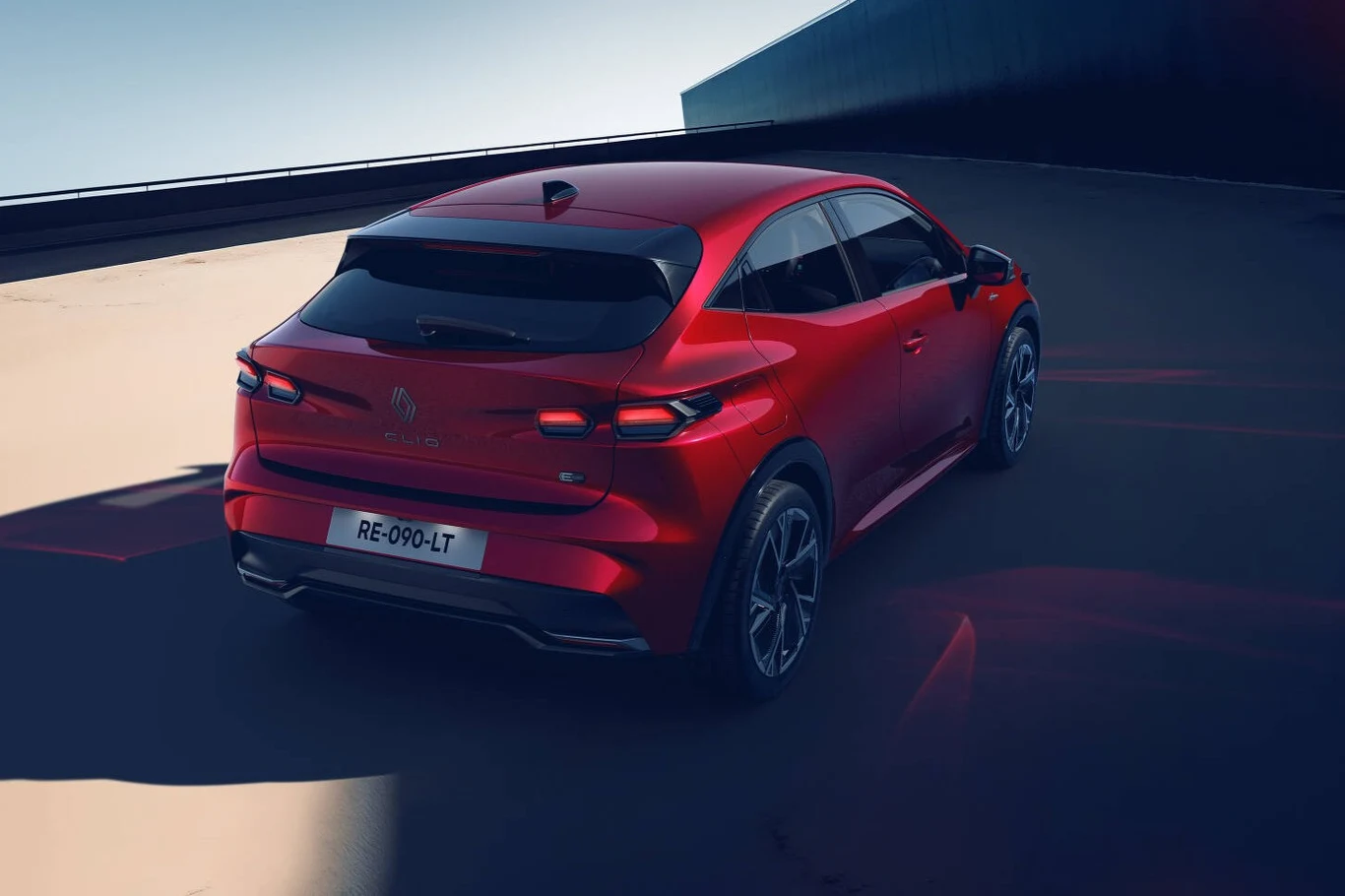 W wariancie hybrydowym Renault Clio zużywa 3,9-4,1 l/100 km. Czerwony, nowoczesny samochód osobowy zaparkowany na pustym, betonowym podłożu, widok od tyłu, z dynamicznymi reflektorami i czarnym dachem oraz szykownymi felgami.