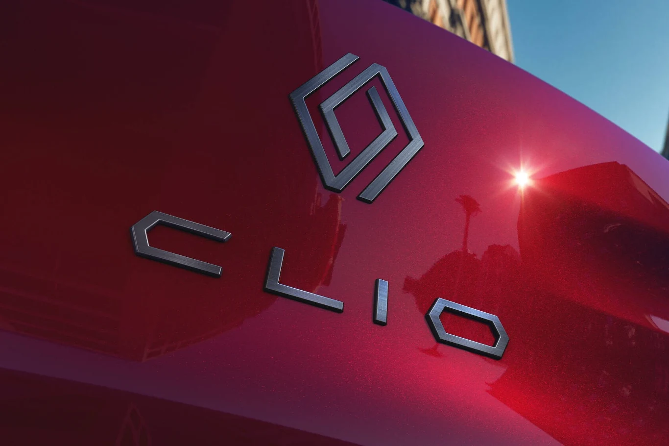 Za nowe Renault Clio trzeba zapłacić co najmniej 84 900 zł. Czerwony lakier samochodu, nowoczesne logo producenta i napis „CLIO” umieszczone na karoserii, odbijająca się sylwetka miasta i promienie słońca.