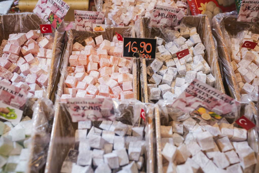 Różnorodne rodzaje lokum tureckiego poukładane w plastikowych pojemnikach, posypane cukrem pudrem, z widocznymi małymi tureckimi flagami i ceną 16,90 za kilogram.