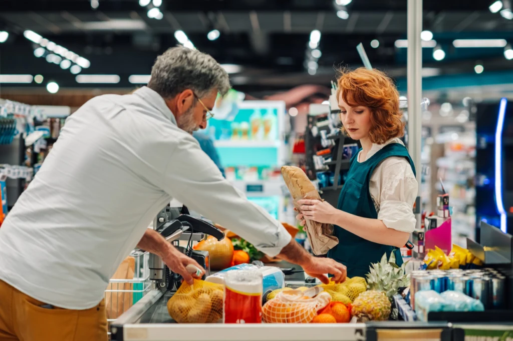 Kasjerka skanuje produkty klienta przy kasie w supermarkecie, na taśmie widoczne są owoce, warzywa, chleb i inne artykuły spożywcze.
