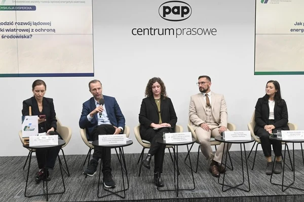 Panel ekspertów siedzących na scenie podczas konferencji prasowej, po środku prowadzący mówi do mikrofonu, reszta osób skupiona, w tle plansze informacyjne oraz logo centrum prasowego.
