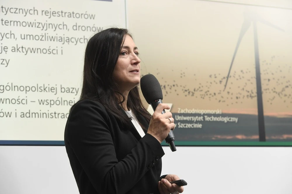 Kobieta w ciemnym żakiecie trzyma mikrofon podczas prezentacji, w tle slajd z tekstem dotyczącym rejestratorów oraz zdjęcie turbiny wiatrowej i logo Zachodniopomorskiego Uniwersytetu Technologicznego w Szczecinie.