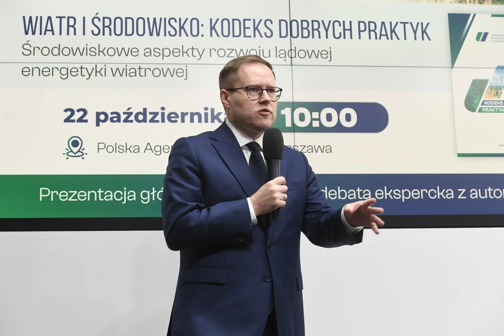 Mężczyzna w okularach i granatowym garniturze przemawia do mikrofonu na tle planszy informacyjnej dotyczącej konferencji o aspekcie środowiskowym energii wiatrowej. W tle napisy oraz logotypy wydarzenia.