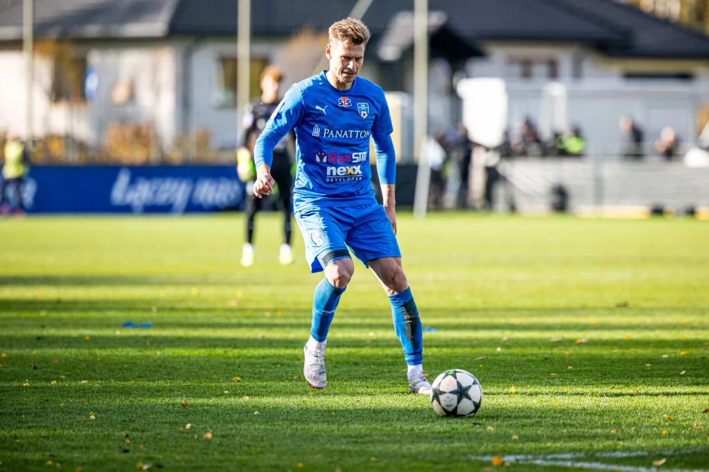 Piłkarz w niebieskim stroju koncentrujący się na prowadzeniu piłki na zielonym boisku, w tle widoczni inni zawodnicy oraz budynki i niewielka grupa kibiców.