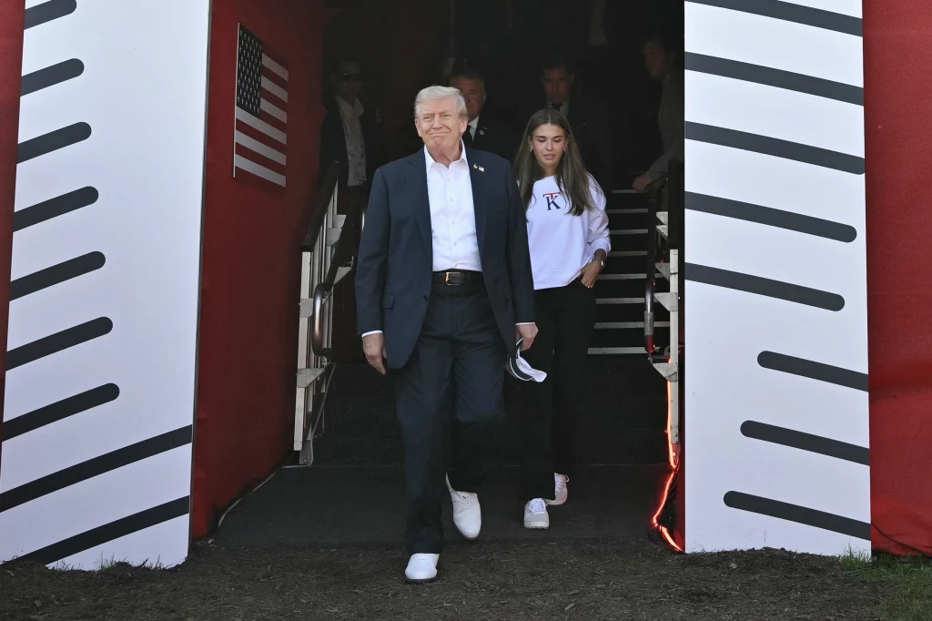 Mężczyzna w garniturze i białych sportowych butach wychodzi z konstrukcji ozdobionej amerykańską flagą, towarzyszy mu młoda kobieta w jasnej bluzie i białych butach. W tle widoczne schody i kilka osób.