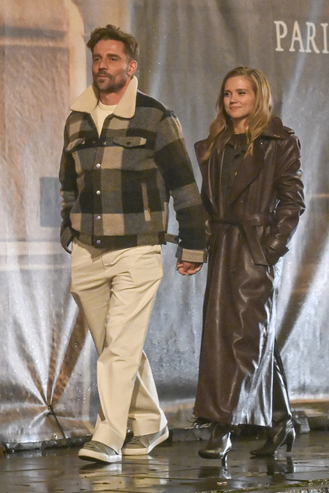 Agnieszka Kaczorowska, Marcin Rogacewicz Para trzymająca się za ręce spaceruje po mieście, kobieta ubrana w długi, brązowy płaszcz i czarne buty, mężczyzna w jasnych spodniach oraz kraciastym płaszczu, oboje wyglądają spokojnie i zrelaksowani.