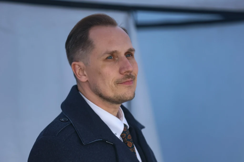 Mężczyzna w średnim wieku ubrany elegancko w białą koszulę, ciemny płaszcz i krawat stoi na tle jasnego, rozmytego tła, patrząc lekko w bok z delikatnym uśmiechem.