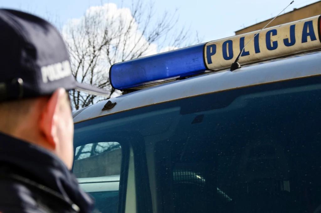 Policjant stojący przy radiowozie z niebieskim kogutem oraz napisem 'Policja', w tle nagie gałęzie drzew i fragment budynku.