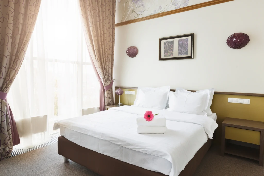 Za noc w hotelu ze śniadaniem zapłacimy obecnie od 450 zł Nowoczesna sypialnia hotelowa z dużym łóżkiem dwuosobowym starannie pościelonym białą pościelą, kwiatem i ręcznikami ułożonymi na środku, obok szerokie okno z zasłonami przepuszczającymi dużo dziennego światła, na ścianie dwa kinkiety i obraz, a obok ł...