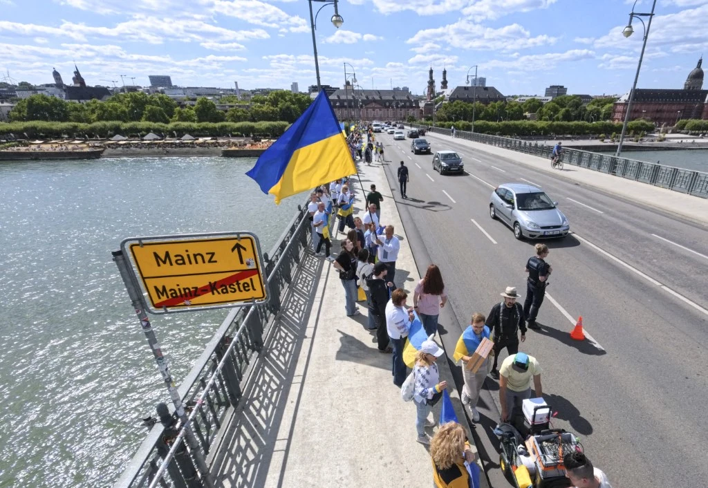 Ukraińcy w Niemczech. Napływ młodych mężczyzn wywołuje protesty prawicowych polityków Ludzie zgromadzeni na moście nad rzeką, trzymający ukraińskie flagi, tworzą rząd osób wzdłuż chodnika. Po drugiej stronie mostu przejeżdżają samochody. Obok widoczny jest znak drogowy z nazwami Mainz i Mainz-Kastel, a w tle zarys miasta.