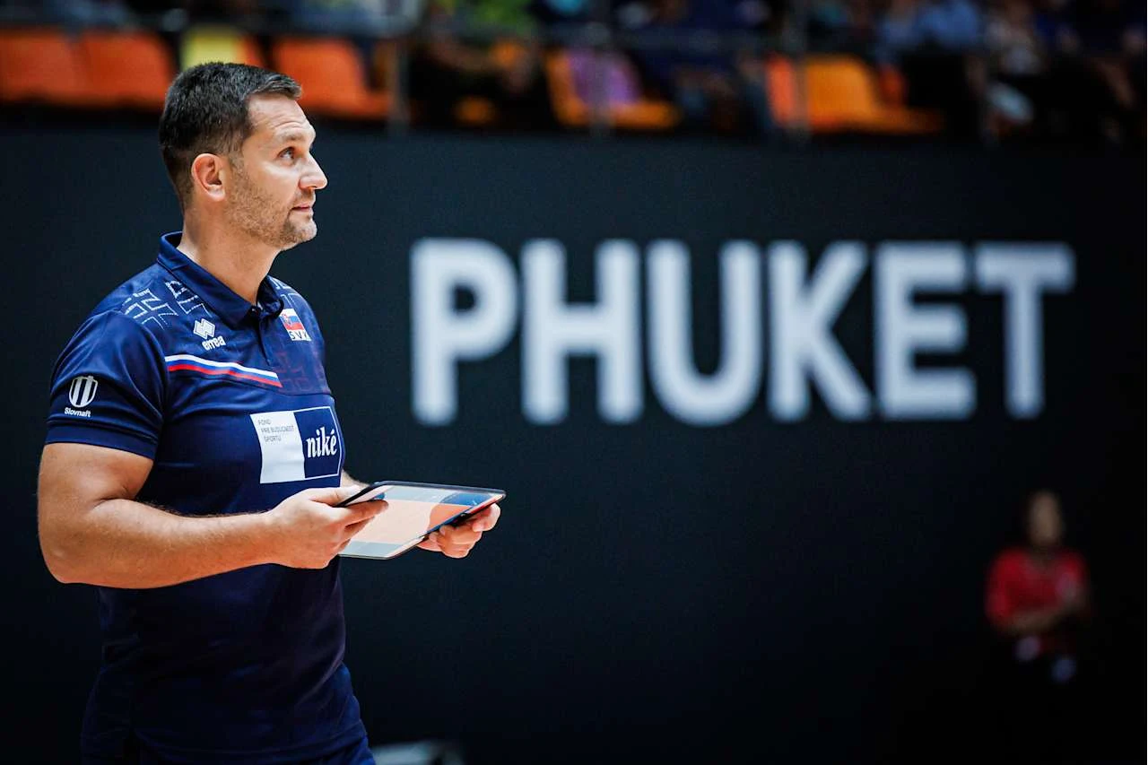 Mężczyzna w sportowej koszulce trzymający tablet stoi na tle dużego napisu PHUKET w hali sportowej.