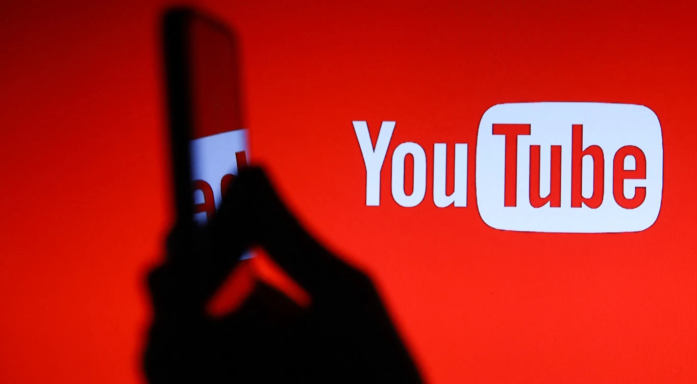 Parodia czy realne zagrożenie? YouTube zdecyduje, czy usunąć deepfake'a. Ale najpierw pozwoli ci go znaleźć swoim narzędziem Logo YouTube na czerwonym tle, czarny zarys dłoni trzymającej telefon na pierwszym planie.