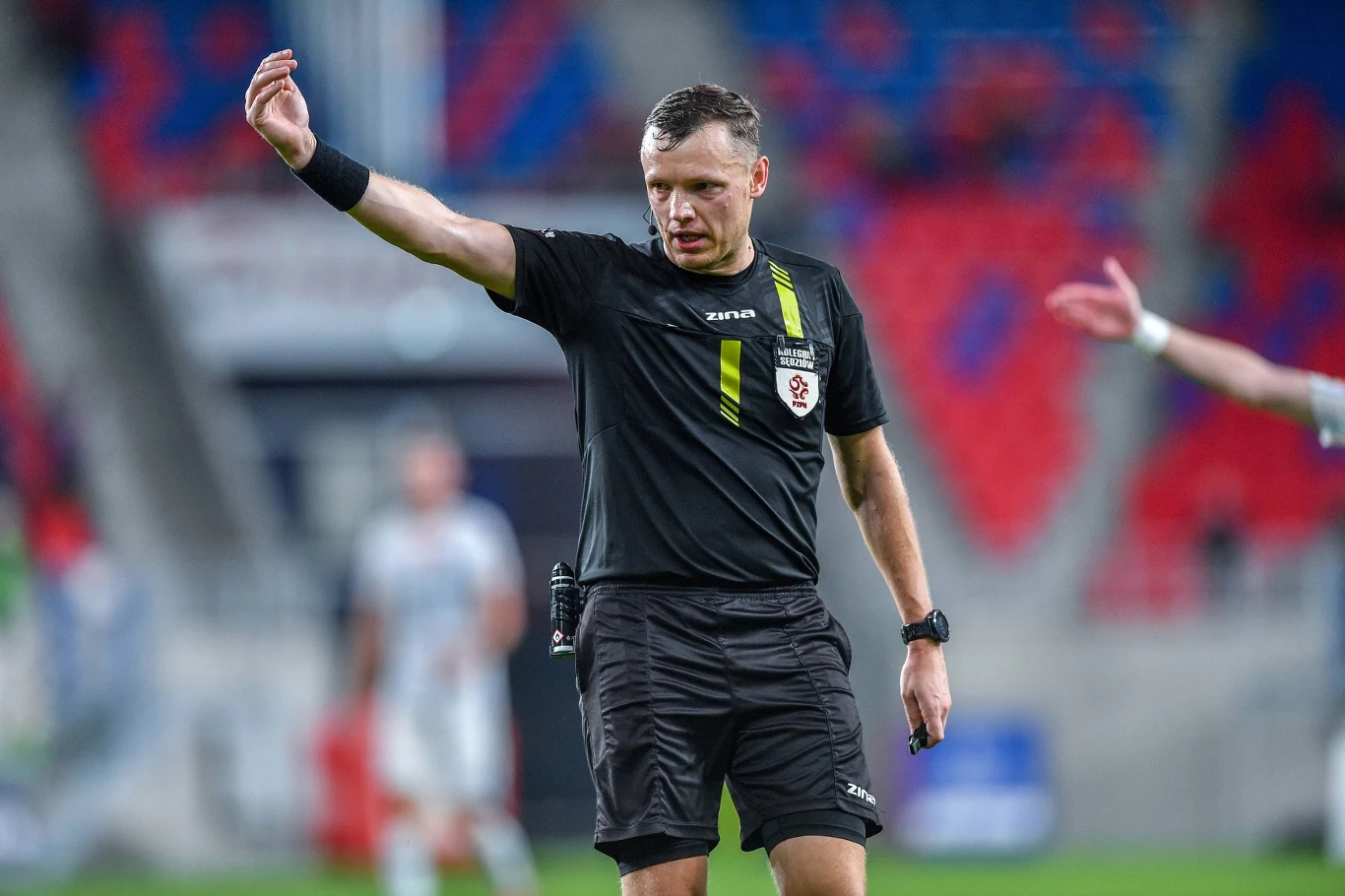 Damian Krumplewski Sędzia piłkarski w czarnym stroju wydaje decyzję podczas meczu piłki nożnej, na tle rozmazanych postaci zawodników oraz trybun stadionowych.