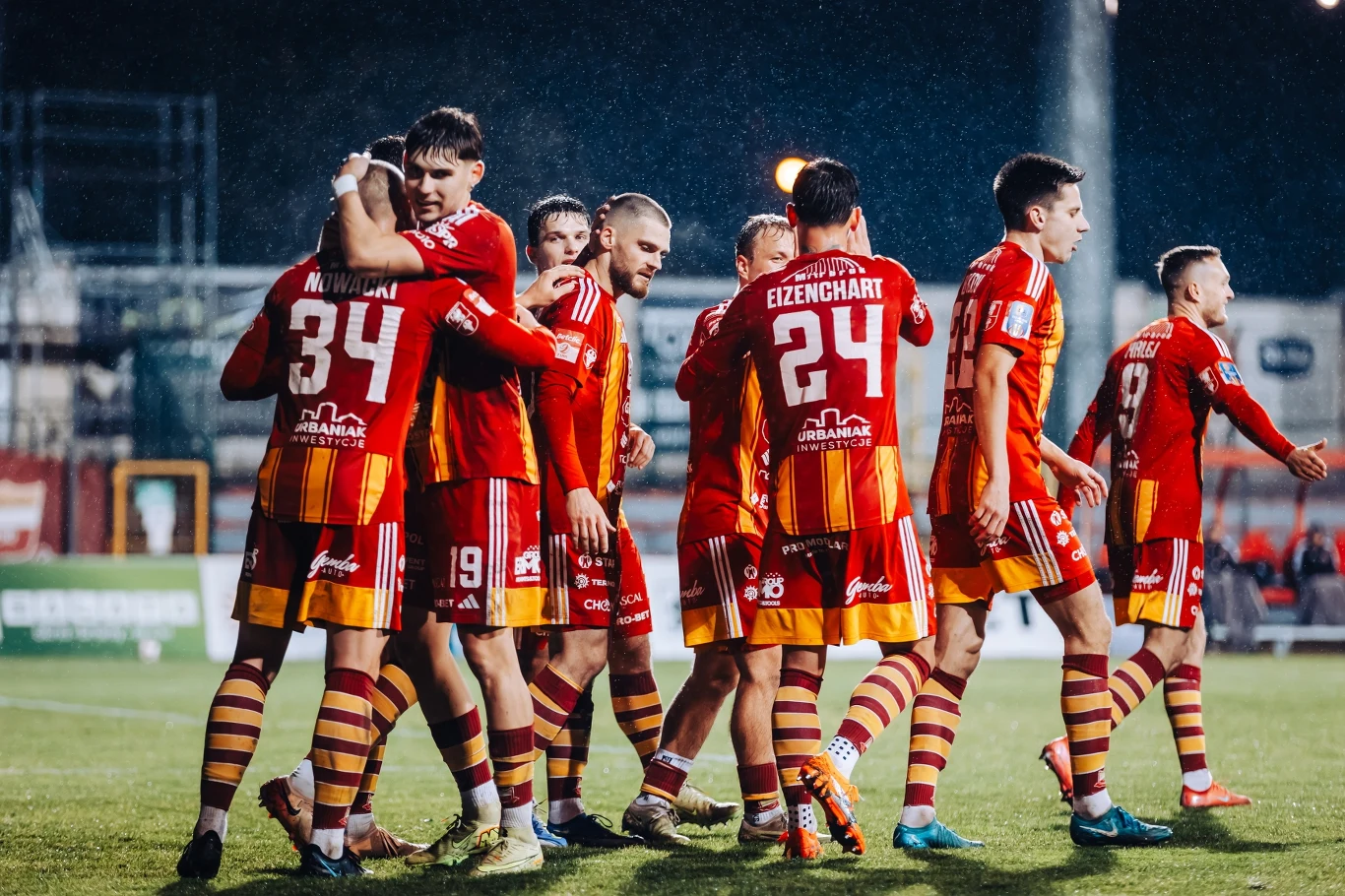 Chojniczanka Chojnice - Świt Szczecin Grupa piłkarzy w czerwono-żółtych strojach celebruje razem na boisku, kilku zawodników się przytula, inni wyrażają radość po udanym meczu, w tle rozmyte trybuny i sztuczne oświetlenie stadionu.