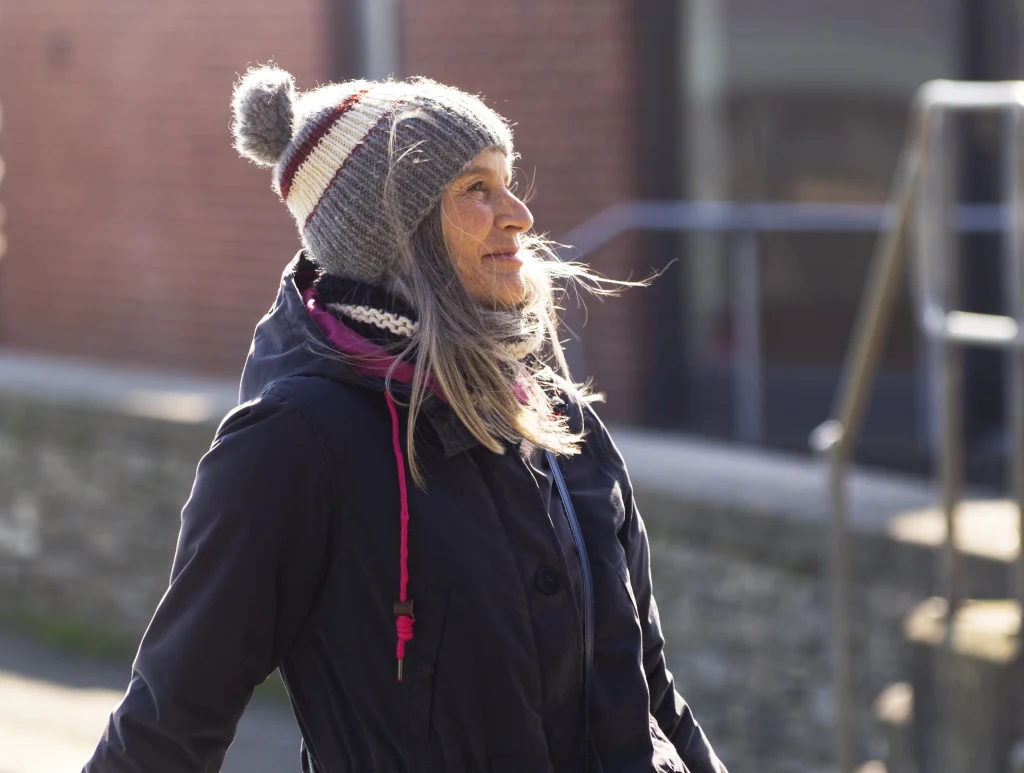 Jakie kolory pasują do siwych włosów? Starsza kobieta z długimi siwymi włosami uśmiecha się i patrzy w bok podczas spaceru na świeżym powietrzu. Ubrana w zimową kurtkę i wełnianą czapkę z pomponem, w tle mur z cegły oraz poręcz.