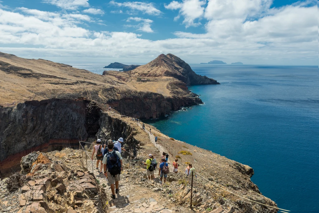 Grupa turystów idąca wzdłuż skalistego klifu z widokiem na ocean, sucha roślinność, błękitne niebo z chmurami i oddalone wyspy na horyzoncie.