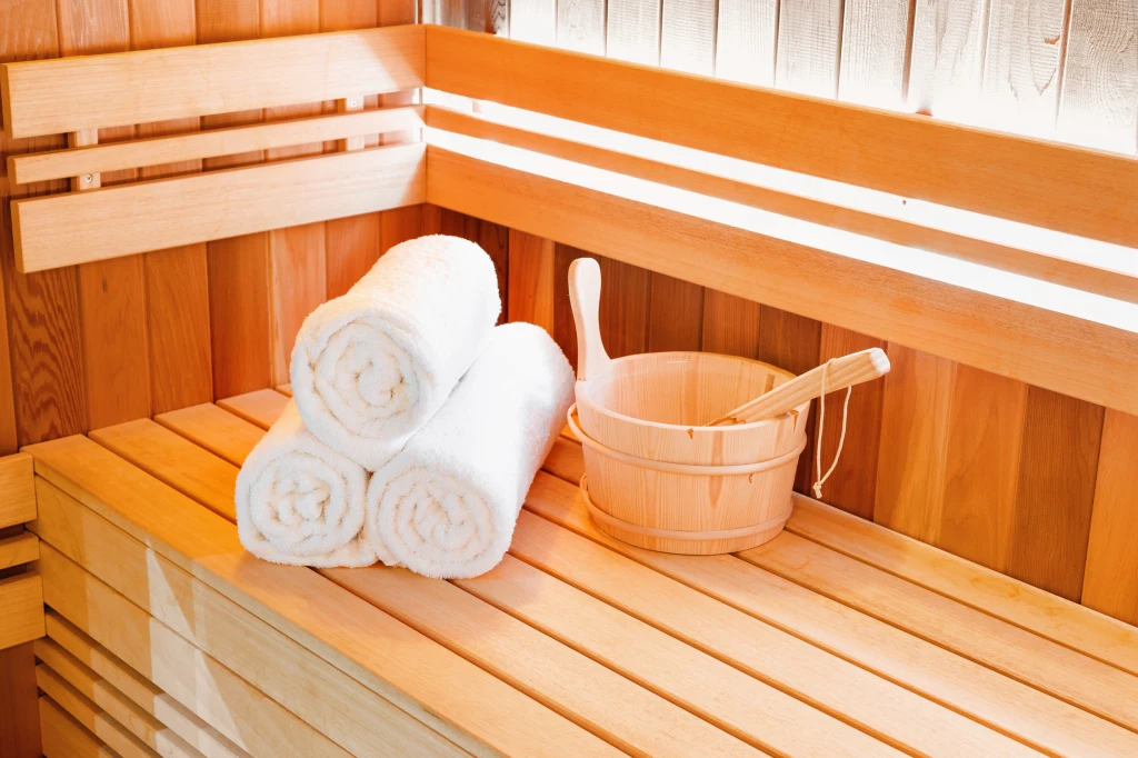Sauna to nie tylko relaks Drewniana ławka w saunie, na której leżą trzy zwinięte w rulony białe ręczniki oraz drewniane wiaderko z drewnianą chochlą, oświetlone naturalnym światłem.