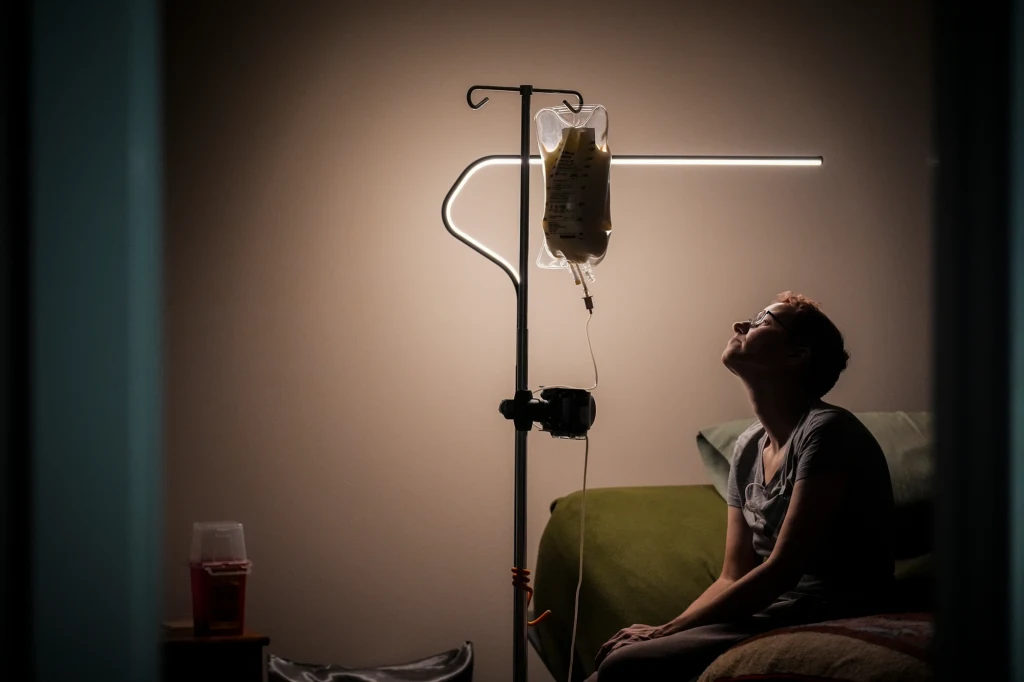 Emily Hoffman śpi ze swoim TPN Osoba siedząca w półmroku na łóżku, podpięta do kroplówki, patrzy w skupieniu na worek z płynem infuzyjnym zawieszony na stojaku. Pokój o stonowanym świetle, w tle łóżko, a na stoliku leży kubek z czerwoną zawartością.
