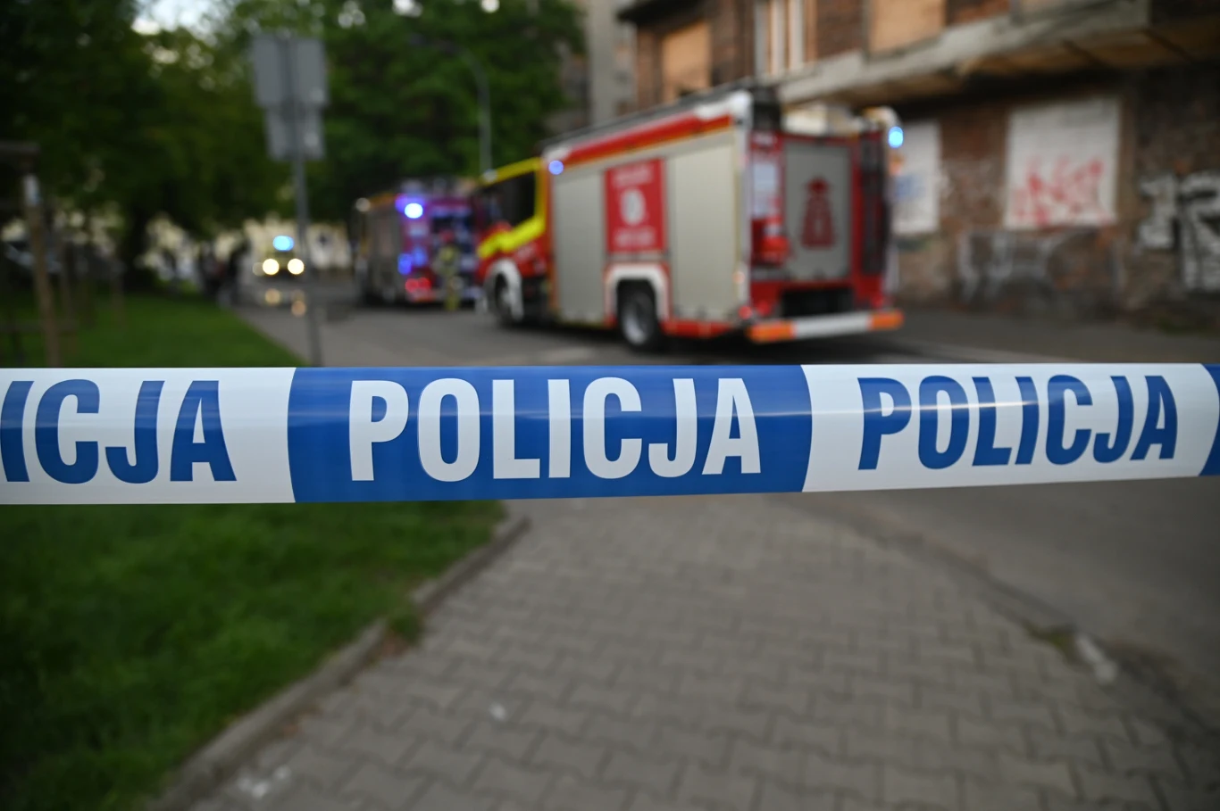 Akcja dziennikarzy magazynu "Interwencja" Polsatu "Sprawiedliwość dla ofiar" rozpoczęta Policyjna taśma odgradzająca teren na miejskim chodniku, w tle samochód straży pożarnej i zniszczony budynek pokryty graffiti.