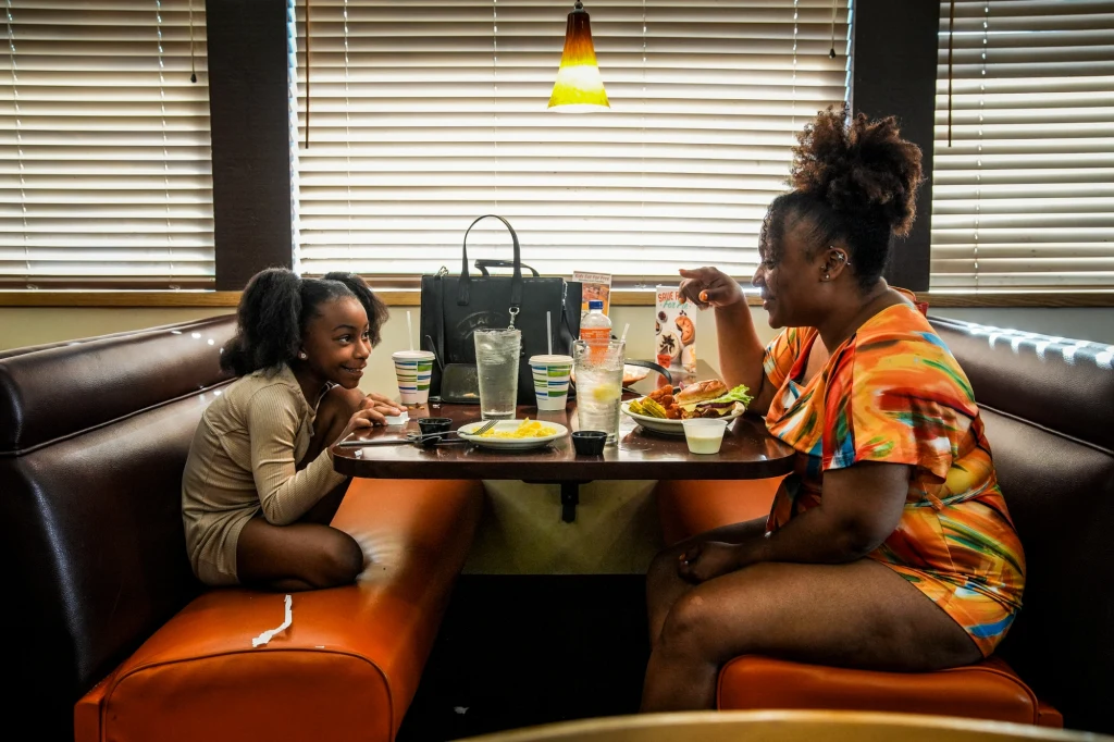 Beatrice Abang-Ntuen ze swoją córką Matka i córka siedzą przy stole w restauracyjnym boksie, uśmiechają się do siebie podczas wspólnego posiłku. Na stole znajdują się talerze z jedzeniem, kubki i szklanki z napojami. Pomiędzy nimi stoi czarna torebka, w tle okno z żaluzjami i lampka sufi...