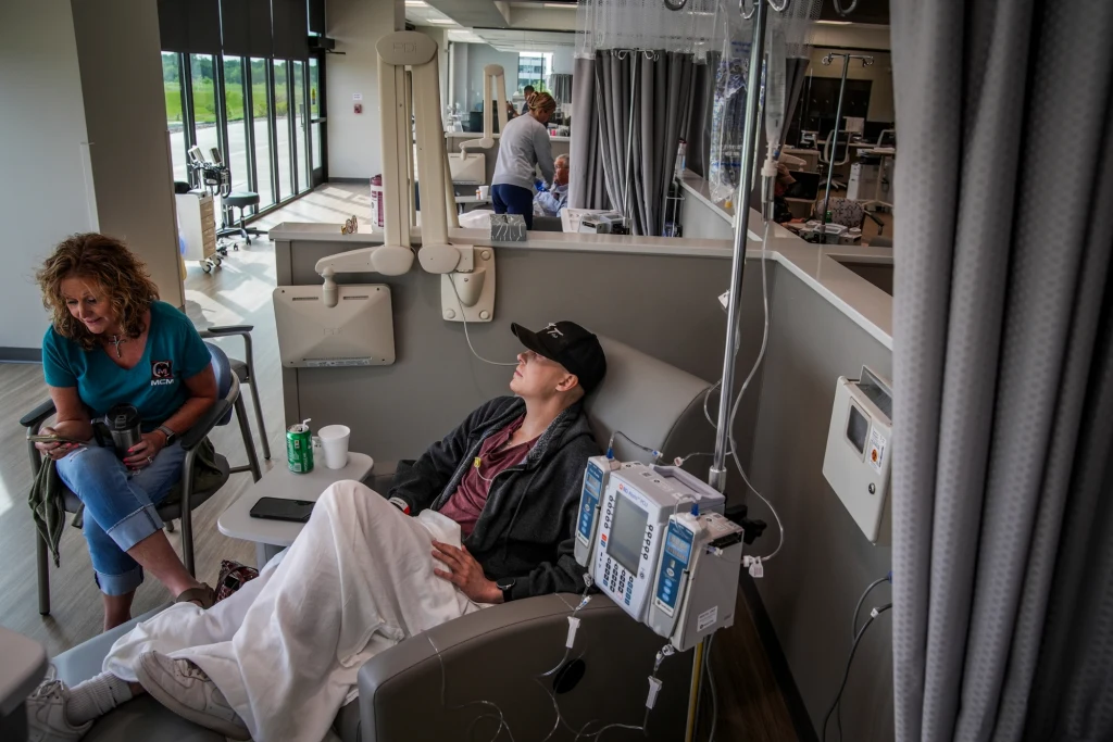 Cole Seiler w szpitalu Młody mężczyzna siedzi na fotelu w szpitalnej sali, przykryty kocem, podłączony do kroplówki, obok niego siedzi kobieta. W tle widoczne są inne stanowiska leczenia oraz medyczny personel zajmujący się pacjentami.