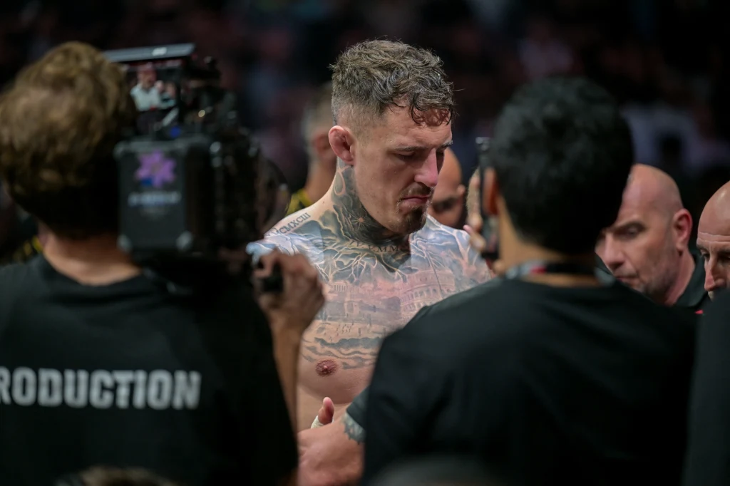 Tom Aspinall Zawodnik z licznymi tatuażami na torsie otoczony przez członków ekipy realizacyjnej oraz operatorów kamer, skupiony, przygotowuje się do wydarzenia sportowego, w tle tłum i rozmyte światła.