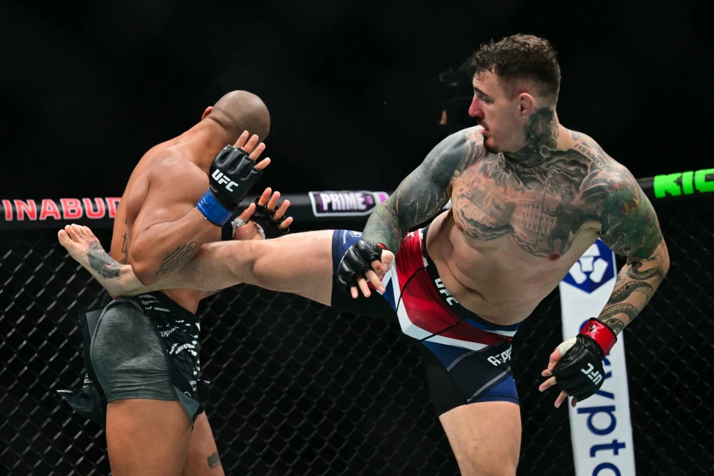 Ciryl Gane i Tom Aspinall w oktagonie Dwóch zawodników MMA w klatce podczas walki, jeden z nich wykonuje kopnięcie w kierunku tułowia rywala, widoczne umięśnione sylwetki i tatuaże, dynamiczna akcja na tle siatki oktagonu.