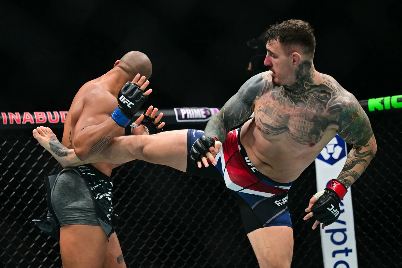 Ciryl Gane i Tom Aspinall w oktagonie Dwóch zawodników MMA w klatce podczas walki, jeden z nich wykonuje kopnięcie w kierunku tułowia rywala, widoczne umięśnione sylwetki i tatuaże, dynamiczna akcja na tle siatki oktagonu.