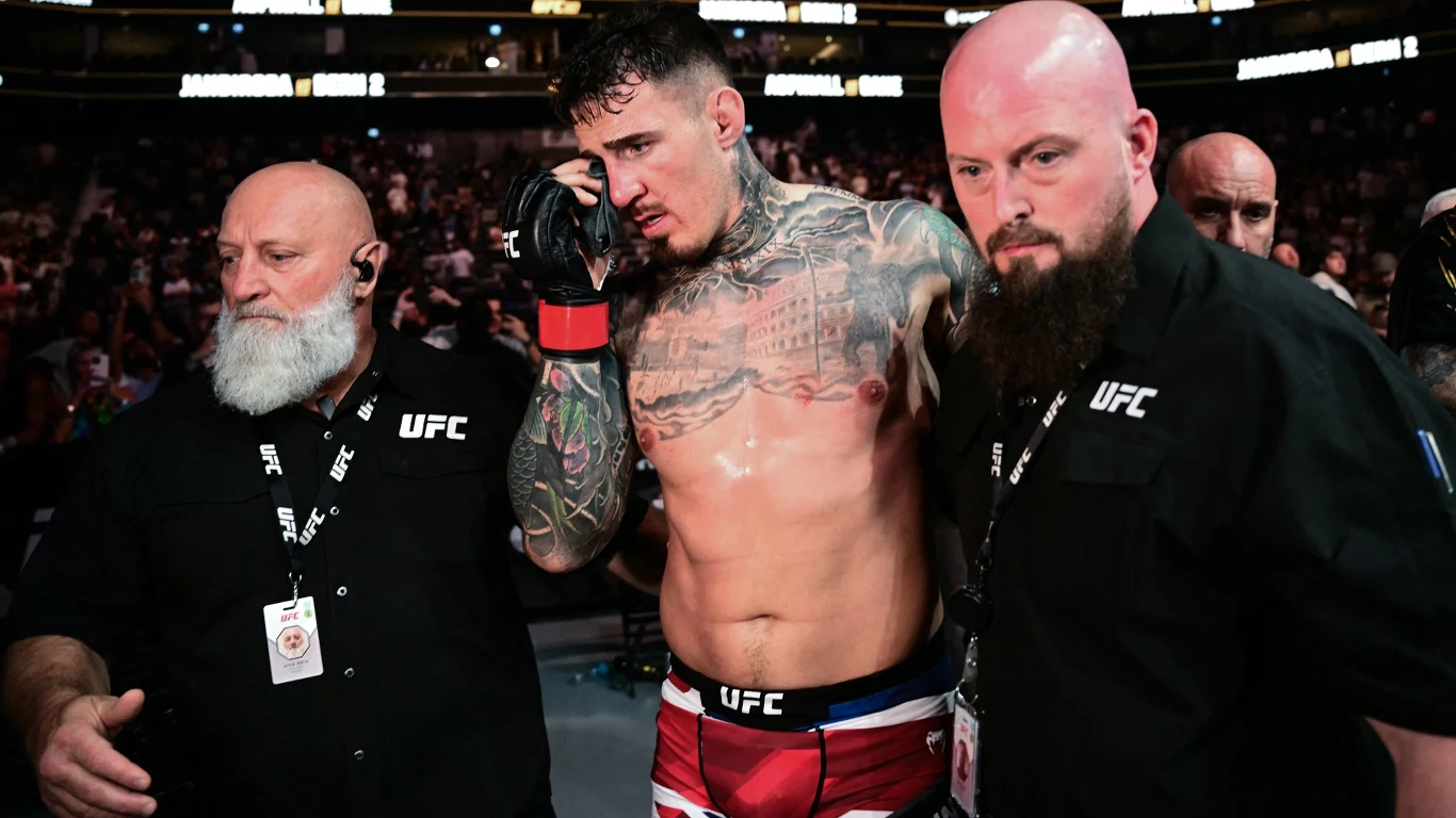 Tom Aspinall ucierpiał w walce z Cirylem Ganem na UFC 321 Zawodnik MMA z licznymi tatuażami wychodzi z oktagonu, otoczony przez dwóch pracowników UFC, wygląda na zmęczonego i poruszonego, trzyma się za twarz, w tle widoczna publiczność na trybunach.