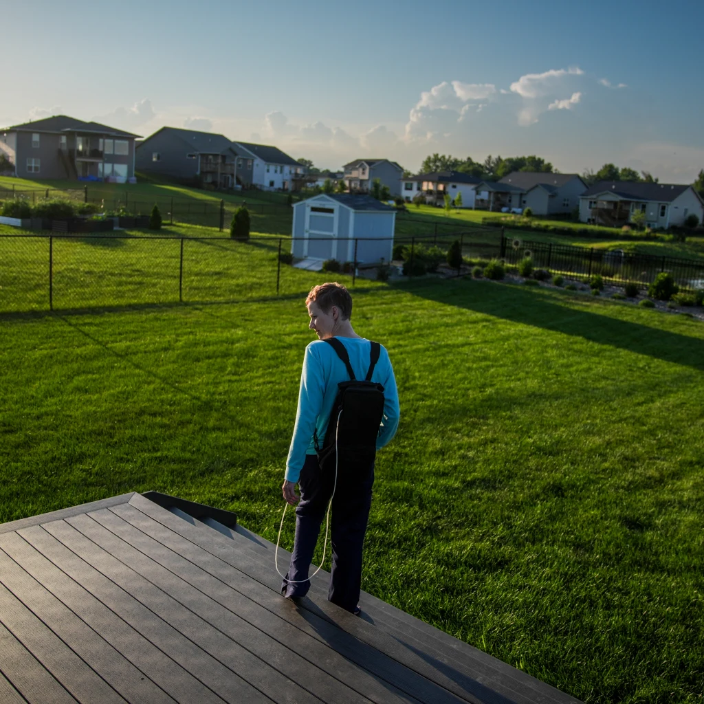 Emily Hoffman musi być karmiona dojelitowo Chłopiec z plecakiem stoi na tarasie skierowany w stronę dużego, zielonego trawnika. W tle widoczne są domy jednorodzinne, porządnie utrzymane ogródki i altanka ogrodowa. Całość otacza ogrodzenie.