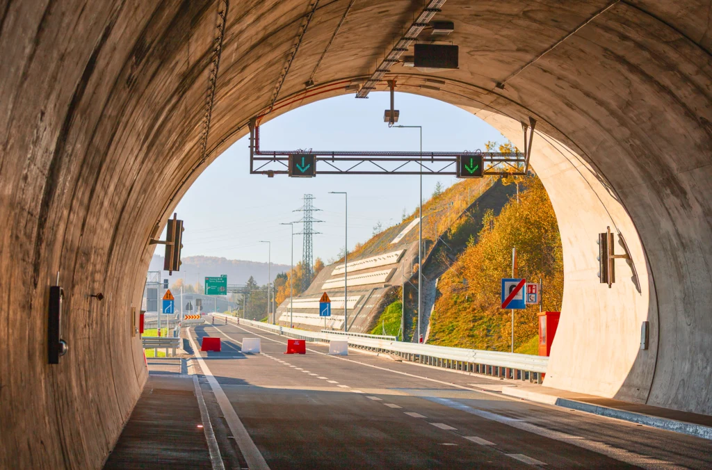Widok wnętrza nowoczesnego tunelu drogowego z wyjazdem na otwartą autostradę, znaki drogowe, zielone strzałki kierunkowe oraz jesienne drzewa na zboczu w tle.