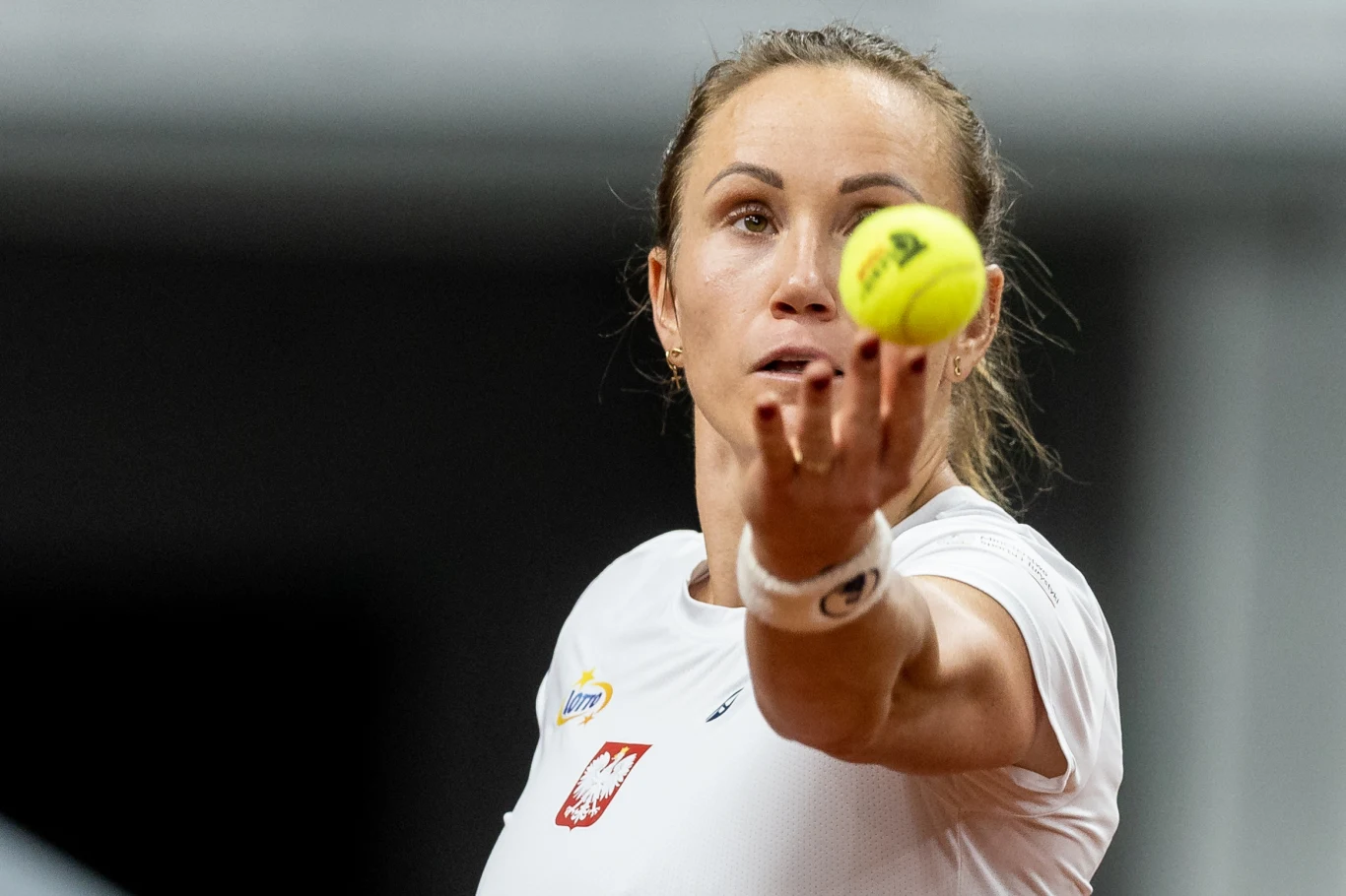 Zawodniczka tenisowa w białej koszulce z polskim godłem w trakcie serwowania piłki na korcie.