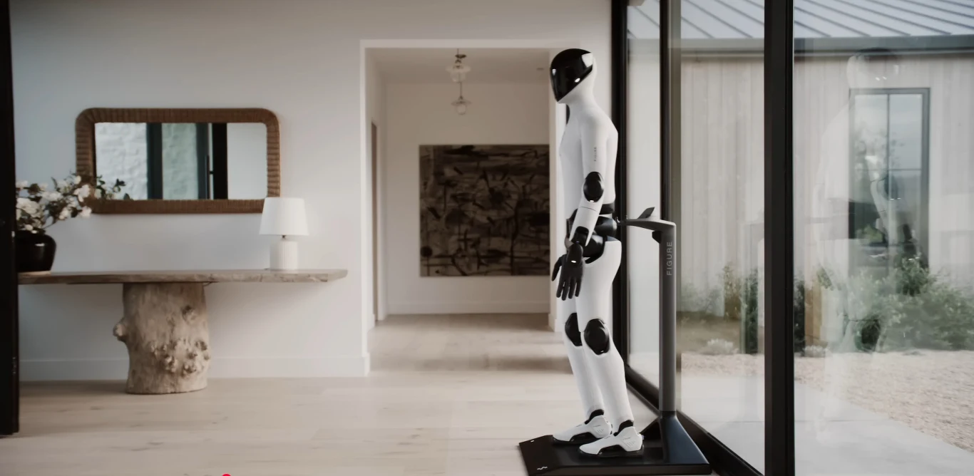 Ładowanie jest bezprzewodowe (indukcja) i przypomina nieco uzupełnianie baterii w odkurzaczach sprzątających Humanoidalny robot stojący w nowoczesnym, jasnym wnętrzu przy dużym oknie, z widokiem na część ogrodu. Obok po lewej stronie znajduje się drewniany stół z lampą i wazonem z kwiatami oraz lustro na ścianie.