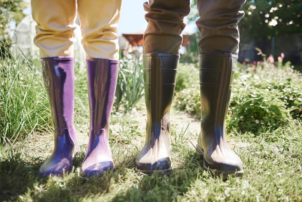 Kalosze chronią przed deszczem, ale ich szczelne tworzywo szkodzi skórze Dwie osoby stojące na trawie w ogrodzie, ubrane w kolorowe, błyszczące kalosze oraz spodnie zakrywające nogi; wokół widoczne zielone rośliny i światło słoneczne.