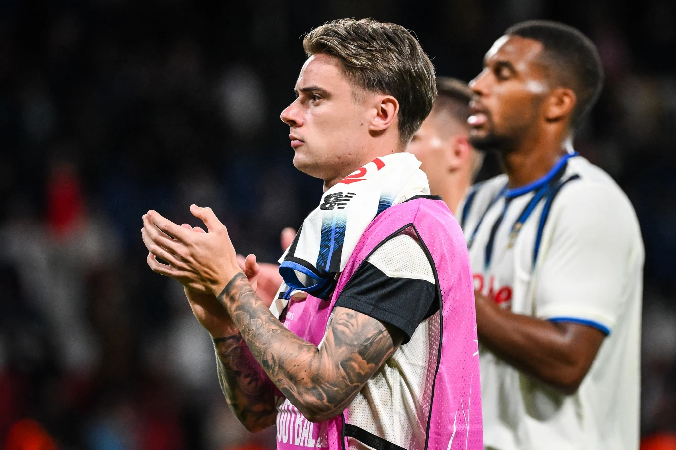 Piłkarz w stroju sportowym klaska na boisku, z ręcznikiem przewieszonym przez ramię, w tle widoczny jest inny zawodnik w białej koszulce. Obaj są skoncentrowani, scena rozgrywa się podczas wydarzenia sportowego, prawdopodobnie meczu piłki nożnej.