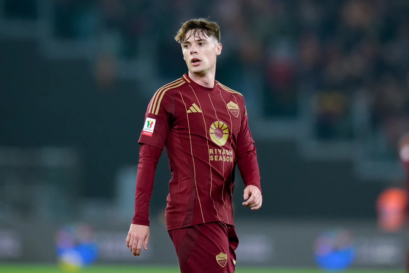 Nicola Zalewski w barwach AS Roma Młody piłkarz w bordowym stroju z herbem klubu oraz złotymi akcentami stoi na boisku podczas meczu, spoglądając w bok. W tle rozmazana publiczność i stadionowe światła.