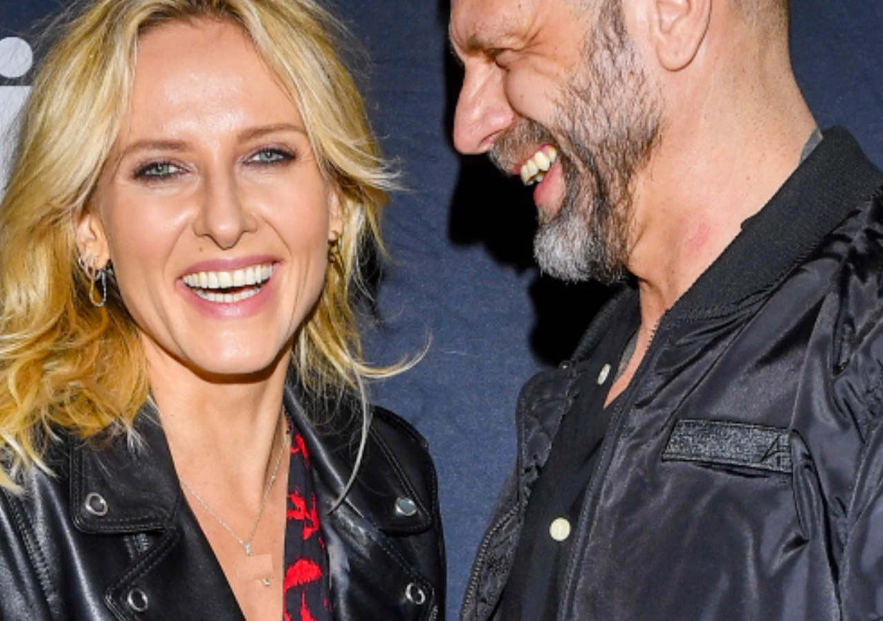 Dwie uśmiechnięte osoby stojące obok siebie na ciemnym tle, kobieta w skórzanej kurtce i sukience w czerwony wzór, mężczyzna w czarnej kurtce, sprawiają wrażenie dobrze bawiących się razem.