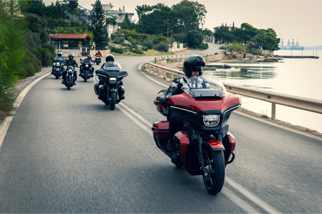 Grupa motocyklistów jadących malowniczą drogą wzdłuż wody, otoczona zielenią i zabudowaniami, prowadzący motocykl w czerwonym kolorze jedzie na czele grupy