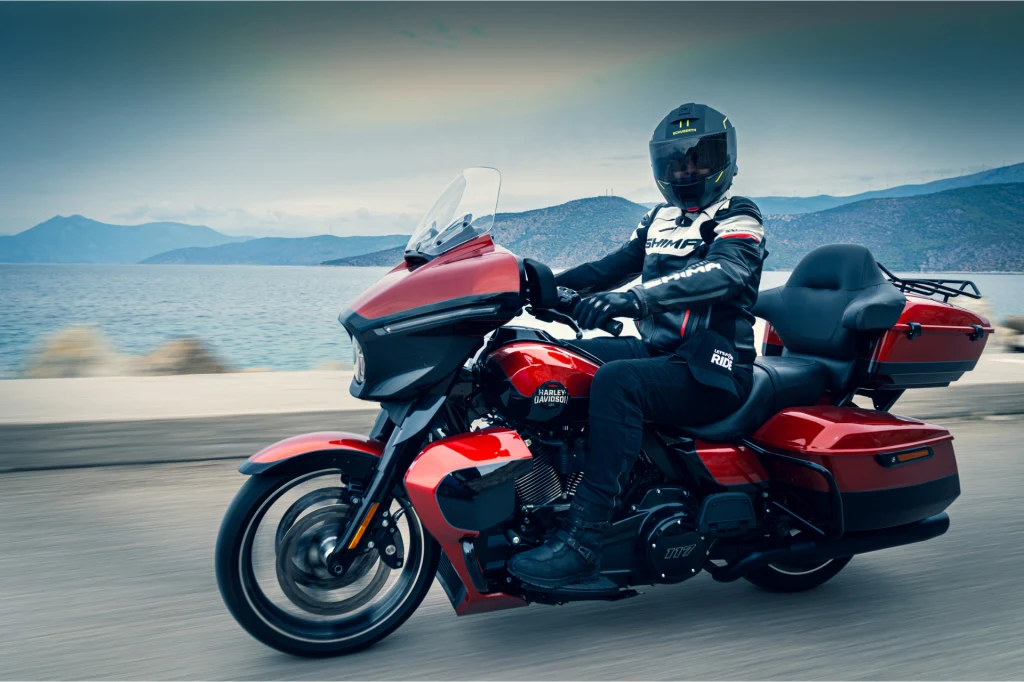 Motocyklista ubrany w strój ochronny jedzie nowoczesnym, czerwonym motocyklem turystycznym wzdłuż drogi znajdującej się nad brzegiem dużego jeziora lub morza, w tle widoczne są góry oraz zachmurzone niebo.