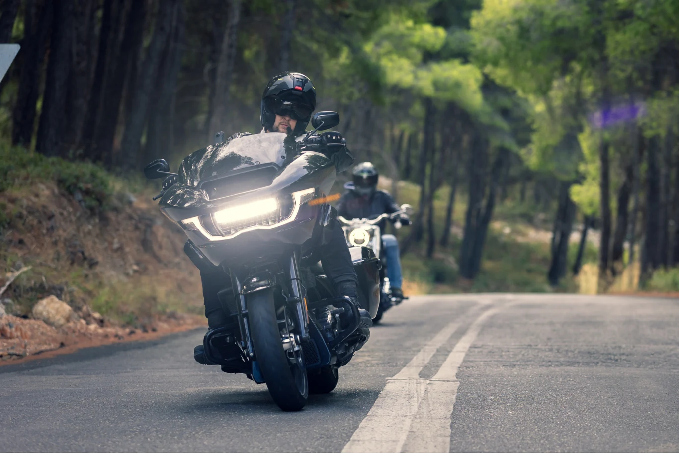 Dwóch motocyklistów w kaskach jedzie asfaltową drogą przez las, prowadząc duże, nowoczesne motocykle. Na pierwszym planie dominują detale maszyny oraz reflektor, w tle rozmyte drzewa i drugi motocyklista.