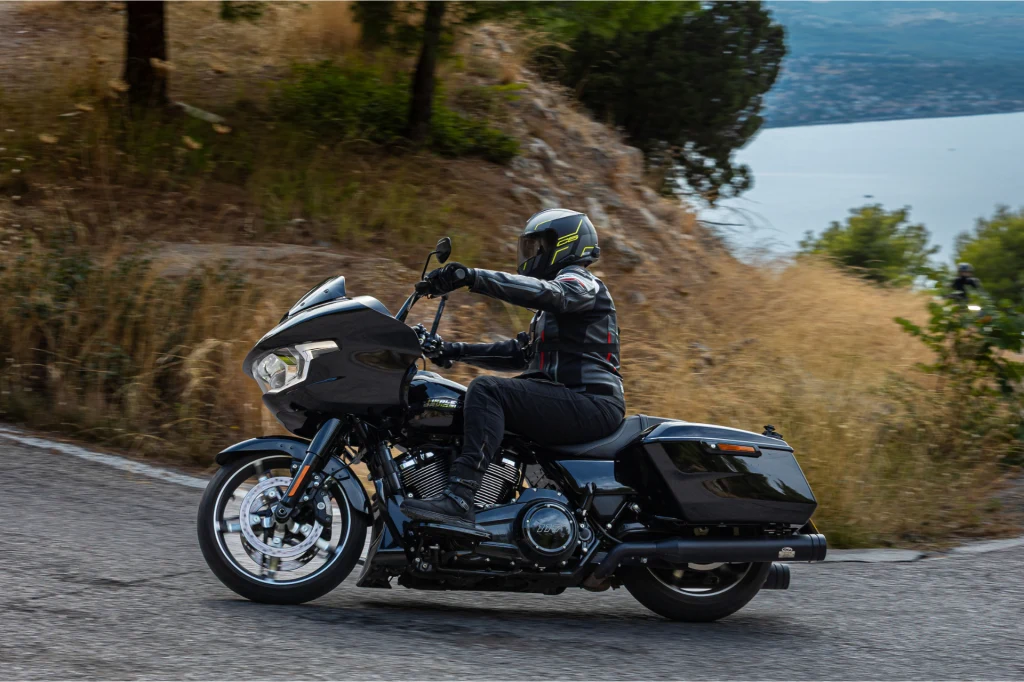 Motocyklista w pełnym stroju ochronnym i kasku jedzie czarnym motocyklem turystycznym po zakręcie asfaltowej drogi w górzystym terenie, w tle widoczne są drzewa, suche trawy oraz niebieska tafla wody.