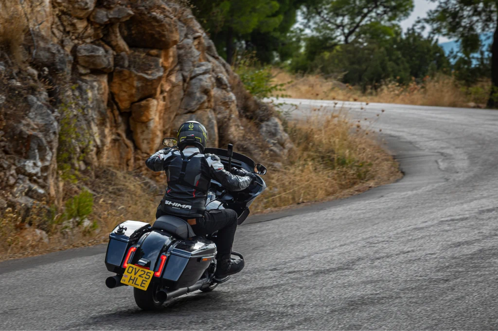Motocyklista w czarnym kombinezonie i kasku pokonuje zakręt na krętej górskiej drodze, otoczonej skałami i suchą roślinnością.