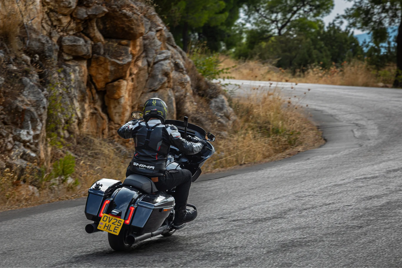 Motocyklista w czarnym kombinezonie i kasku pokonuje zakręt na krętej górskiej drodze, otoczonej skałami i suchą roślinnością.